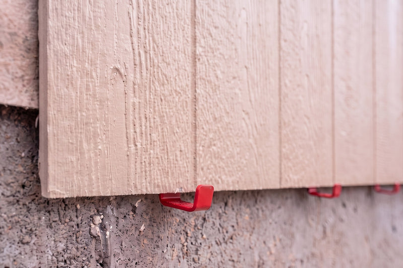 A siding panel bottom edge resting on a row of red EZpanl hooks nailed to the wall
